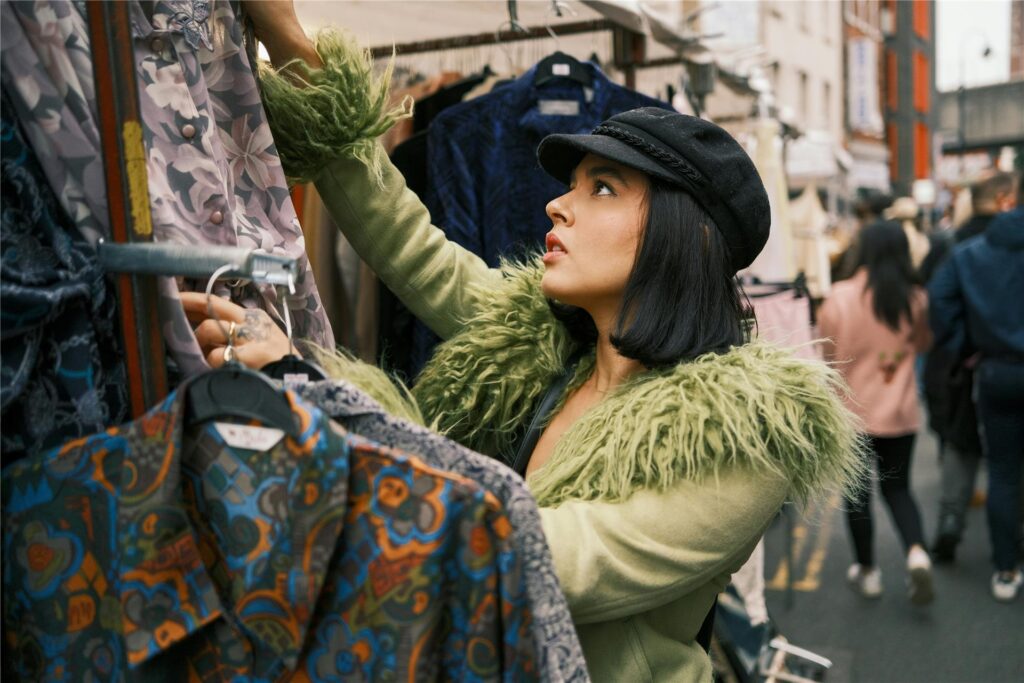 Eine Frau mit grüner Jacke und schwarzer Kappe durchstöbert auf einem Flohmarkt einen Stand mit Blusen und Hemden. Eine Frau mit grüner Jacke und schwarzer Kappe durchstöbert auf einem Flohmarkt einen Stand mit Blusen und Hemden.