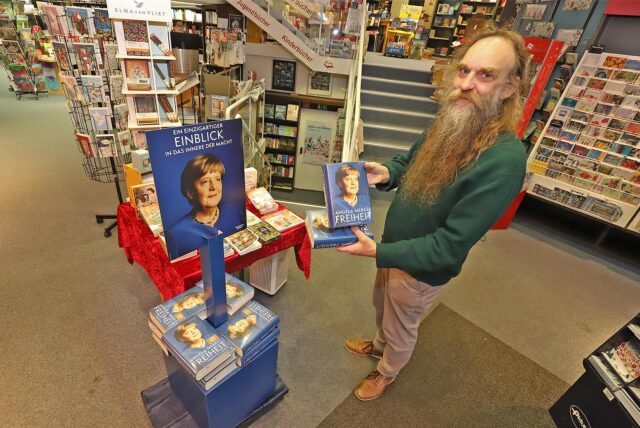 Gordan Friese ist Inhaber der Buchhandlung G. Hornung. Er trägt einen langen Bart, einen grünen Pullover und eine helle Hose. Er hält das neue Buch „Freiheit“ von Angela Merkel in der Hand.