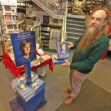 Gordan Friese ist Inhaber der Buchhandlung G. Hornung. Er trägt einen langen Bart, einen grünen Pullover und eine helle Hose. Er hält das neue Buch „Freiheit“ von Angela Merkel in der Hand.