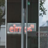 Das Logo des Unternehmens Elmos ist durch eine automatische Schiebetür zu sehen.