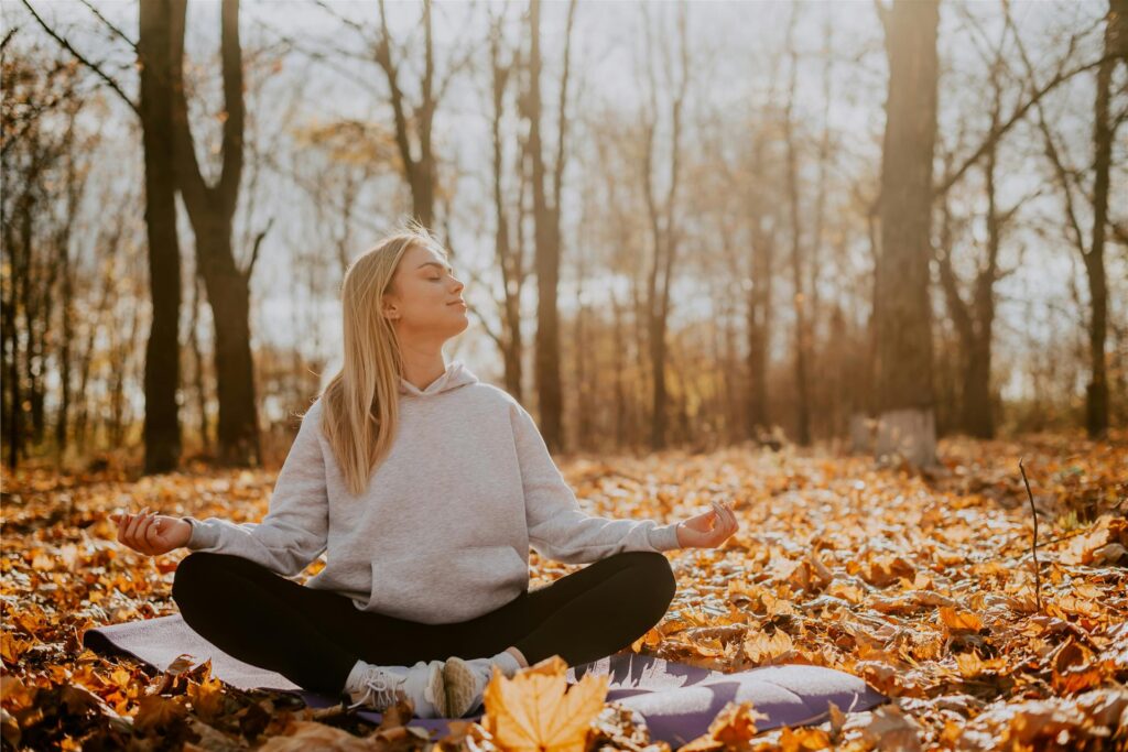 Eine Frau sitzt im Herbst im Wald auf einer Yoga-Matte und meditiert. Mit geschlossenen Augen hält sie ihr Gesicht in die Sonne.