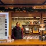Maria Remenski, Inhaberin von „Die Weinhütte“, steht in ihrer Bude. Sie trägt eine schwarze Weste und eine burgunderrote Fleecejacke. Neben ihr ist die Getränkekarte zu sehen.