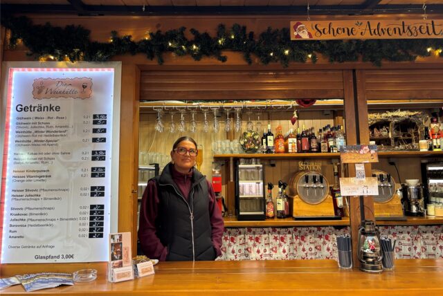 Maria Remenski, Inhaberin von „Die Weinhütte“, steht in ihrer Bude. Sie trägt eine schwarze Weste und eine burgunderrote Fleecejacke. Neben ihr ist die Getränkekarte zu sehen.
