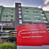Vor dem Eingang vom Hellmig-Krankenhaus in Kamen steht ein rotes Schild mit dem Logo des Krankenhauses.