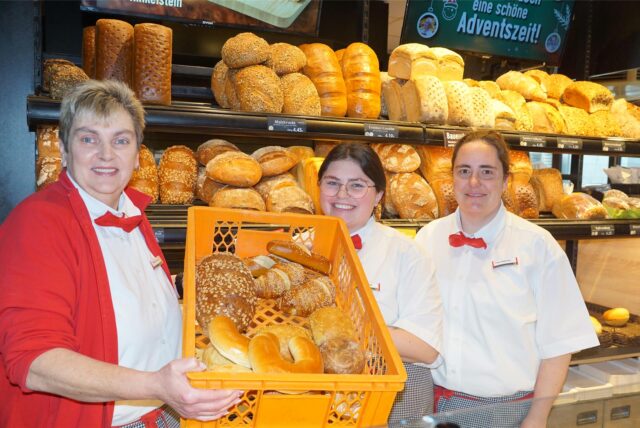 Drei Bäckereifachverkäuferinnen stehen im Verkaufsraum und lächeln. Sie halten einen Korb mit Brötchen in die Kamera.