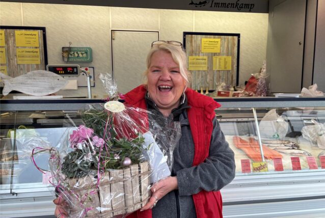 Susanne „Fisch-Susi“ Krenzin-Dickheiwer trägt an ihrem Abschiedstag eine rote Weste und eine graue Fleecejacke. In den Händen hält sie einen Geschenkkekorb mit Blumen.
