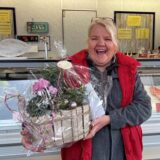 Susanne „Fisch-Susi“ Krenzin-Dickheiwer trägt an ihrem Abschiedstag eine rote Weste und eine graue Fleecejacke. In den Händen hält sie einen Geschenkkekorb mit Blumen.