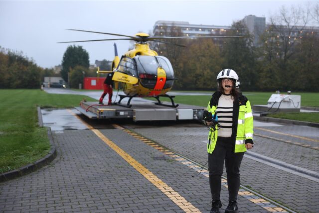 Vor einem gelben Hubschrauber stehen zwei Menschen und reden. Eine Person hält ein Mikrofon.