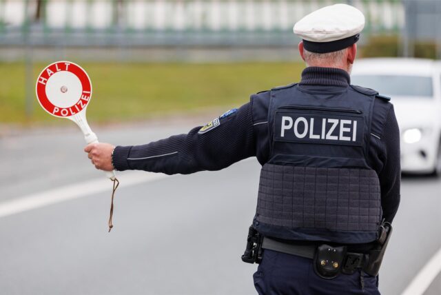 Ein Polizist in Uniform wird von hinten gezeigt. Er streckt eine weiß-rote Kelle raus. Auf der Kelle steht „Halt Polizei“ geschrieben.