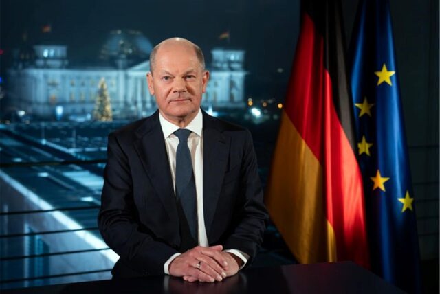 Bundeskanzler Olaf Scholz (SPD) sitzt während eines Fototermins nach der Aufzeichnung seiner Neujahrsansprache im Kanzleramt vor der Kulisse des von Scheinwerfern angestrahlten Reichstages.