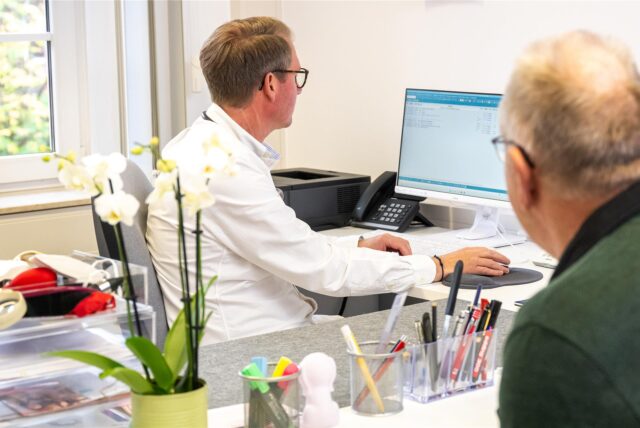 Ein Arzt sitzt an einem Computer, am Tresen in der Praxis steht ein älterer Patient.