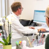 Ein Arzt sitzt an einem Computer, am Tresen in der Praxis steht ein älterer Patient.