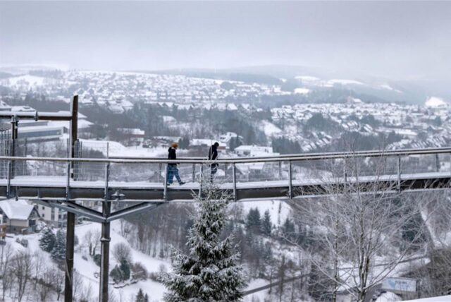 Spaziergänger gehen über den Skywalk neben der Bobbahn mit Blick auf die Landschaft im Schnee.