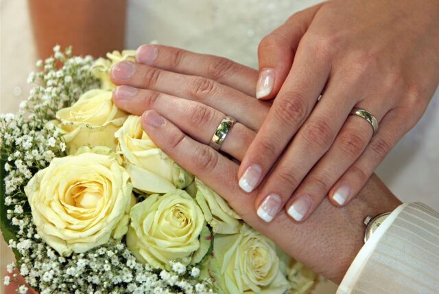 Ein frisch vermähltes Hochzeitspaar hält die Hände mit den Trauringen über dem Brautstrauß.