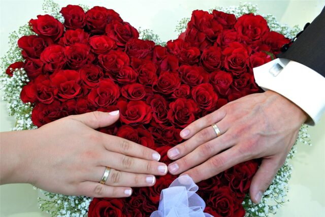Ein Hochzeitspaar hält seine Hände mit den Ringen über ein Herz aus roten Rosen.
