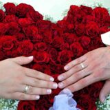 Ein Hochzeitspaar hält seine Hände mit den Ringen über ein Herz aus roten Rosen.