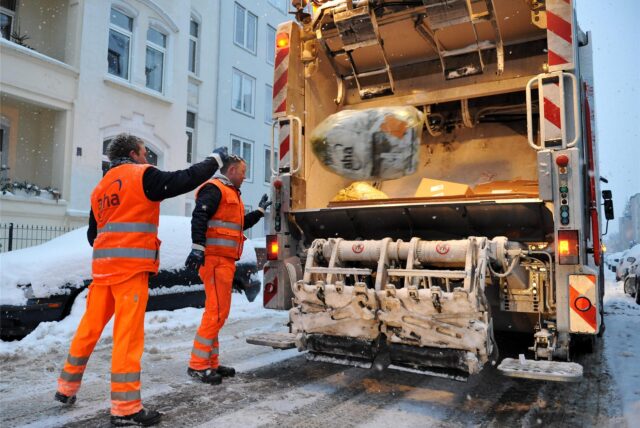 Zwei Müllwerker werfen Müllsäcke in ein Müllfahrzeug.