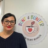 Tanja Au steht in einer Tür auf der ein Logo mit der Aufschrift „Bunt & Bunter Keramik selbst bemalen“ prangt.