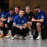 Handballspieler sitzen auf einer Bank. Vor ihnen kniet ihr Trainer und ist mit einem von ihnen im Austausch.