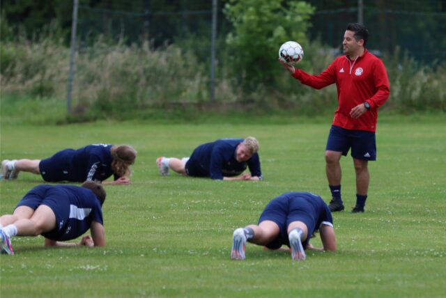 Kurtulus Öztürk hält einen Ball, unter ihm machen Fußballer Liegestütze.