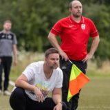 Ein Trainer hockt neben seinen Assistenten am Spielfeldrand und beobachtet mit kritischem Blick ein Fußballspiel.
