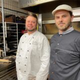 Benjamin Brasse (links) und Marcel Appel grinsen in die Kamera. Brasse trägt sein Bäcker-Outfit, Appel seine Koch-Gewand.