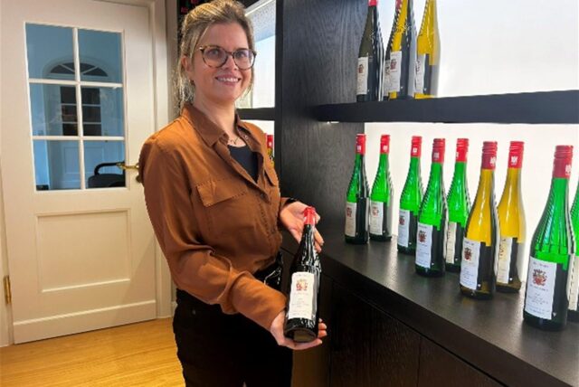 Anita Brandl steht vor einem Weinregal und hält eine Flasche Wein in die