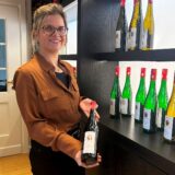 Anita Brandl steht vor einem Weinregal und hält eine Flasche Wein in die