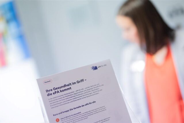 Ein Flyer zur ePA wird vor die Kamera gehalten, dahinter ist unscharf eine Frau in weißem Kittel zu erkennen.