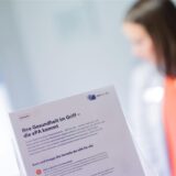 Ein Flyer zur ePA wird vor die Kamera gehalten, dahinter ist unscharf eine Frau in weißem Kittel zu erkennen.