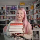 Die Booktok-Influencerin Nathalie Bröske sitzt in ihrem Lesezimmer mit vielen Bücherregalen und hält vier Bücher in die Kamera.