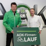 Jörg Kock (AOK-Serviceregionsleiter) und Marcus Hoselmann (upletics GmbH) halten ein Schild des AOK-Firmenlaufs hoch.