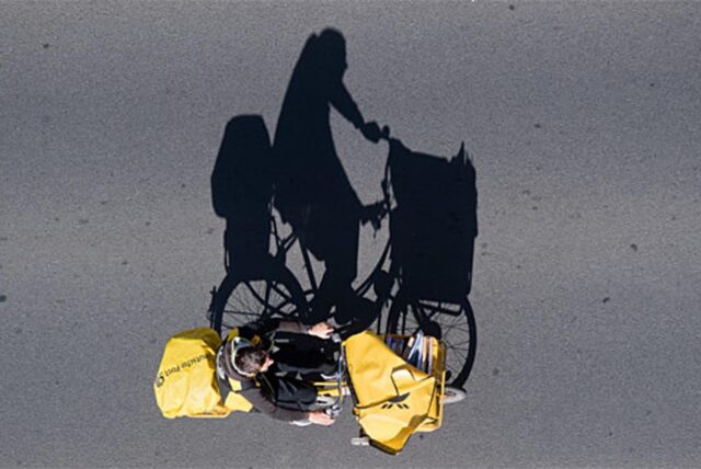 Ein Postbote auf einem Fahrrad wirft einen langen Schatten auf eine Straße.
