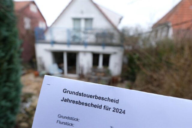 In Brief mit der Aufschrift Grundsteuerbescheid wird in die Kamera gehalten. Im Hintergrund ist unscharf ein Wohnhaus zu erkennen.