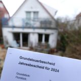 In Brief mit der Aufschrift Grundsteuerbescheid wird in die Kamera gehalten. Im Hintergrund ist unscharf ein Wohnhaus zu erkennen.