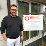 Der Orthopäde und Unfallchirurg Dr. Akin Uzun steht mit einem schwarzen Pullover und einer weißen Hose vor dem Eingangsschild seiner neuen Praxis auf der Allee 3 in Holzwickede.