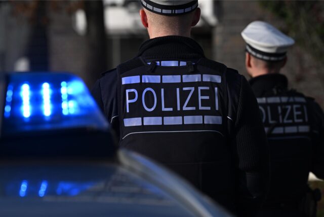 Das Logo der Polizei auf der Dienstkleidung von Polizisten die vor einem Blaulicht stehen