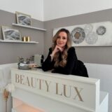 Olga Saprin-Maisinger, Inhaberin von Beauty Lux in Holzwickede. Sie steht hinter ihrem weißen Empfang mit dem goldenen Logo, trägt ein schwarzes Kleid und eine silberne Uhr.