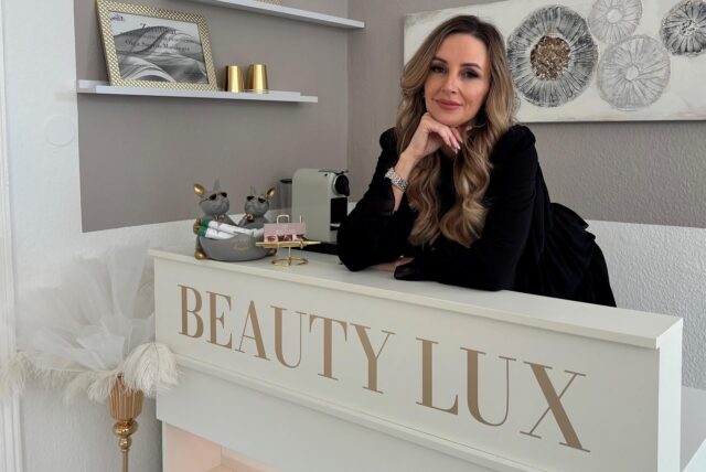Olga Saprin-Maisinger, Inhaberin von Beauty Lux in Holzwickede. Sie steht hinter ihrem weißen Empfang mit dem goldenen Logo, trägt ein schwarzes Kleid und eine silberne Uhr.