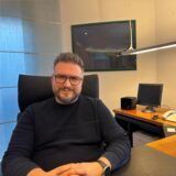 Dr. Uzun ist der neue Orthopäde und Unfallchirurg in Holzwickede. Hier sitzt er in seinem Büro auf der Allee 3. Er trägt eine Brille, einen dunklen Pullover und eine Uhr. Im Hintergrund ist ein Bilderrahmen sowie ein Bildschirm und ein Telefon.