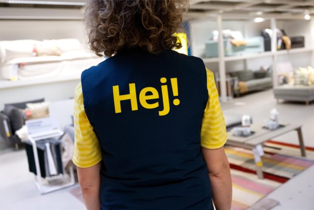 Eine Ikea-Mitarbeiterin im Ikea mit ihrer Arbeitskleidung von hinten fotografiert. Auf dem Rücken steht die Aufschrift „Hej!“.