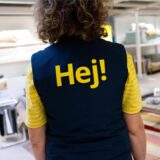 Eine Ikea-Mitarbeiterin im Ikea mit ihrer Arbeitskleidung von hinten fotografiert. Auf dem Rücken steht die Aufschrift „Hej!“.
