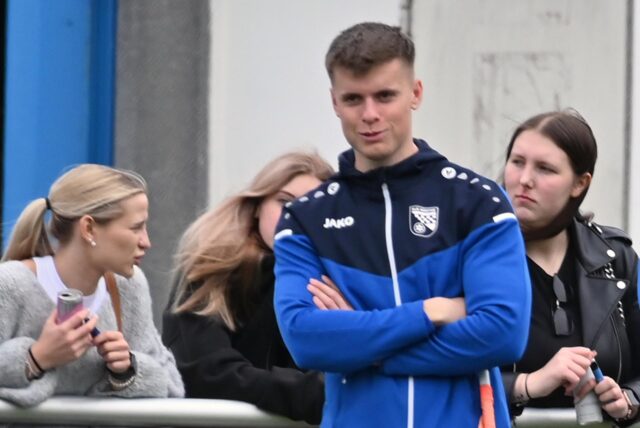 Rünthes Co-Trainer Niklas Barks (M.) steht mit verschränkten Armen an der Bande. Hinter ihm unterhalten sich weibliche Fans.