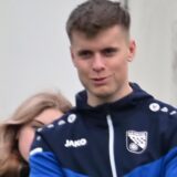 Rünthes Co-Trainer Niklas Barks (M.) steht mit verschränkten Armen an der Bande. Hinter ihm unterhalten sich weibliche Fans.