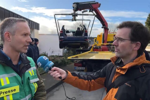 Kevin Kohues (l.), Pressesprecher der Stadt Unna, spricht mit Reporter Thomas Raulf. Im Hintergrund wird ein Autowrack auf einen Abschleppwagen geladen.
