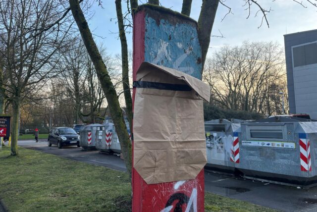 Ein rotes Holzschild, worauf sich schon Moos gebildet hat, mit einer Papiertüte verkleidet. Darunter ein SA-Schriftzug.