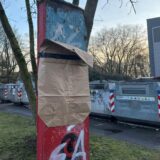 Ein rotes Holzschild, worauf sich schon Moos gebildet hat, mit einer Papiertüte verkleidet. Darunter ein SA-Schriftzug.