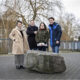 Vier Menschen stehen hinter einem Stein, der auf einer Brücke liegt.