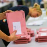 Zwei Hände sortieren rosa Briefwahlumschläge. Im Hintergrund ist eine Frau unscharf zu erkennen, die ebenfalls Wahlunterlagen sortiert.