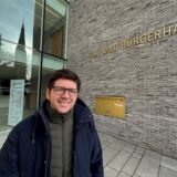 Stefan Thiel ist der neue Fachbereichsleiter der Wirtschaftsförderung der Gemeinde Holzwickede. Hier steht er in eine blauen Jacke und einer grünen Steppweste vor dem Rat und Bürgerhaus in Holzwickede. Thiel trägt eine Brille.